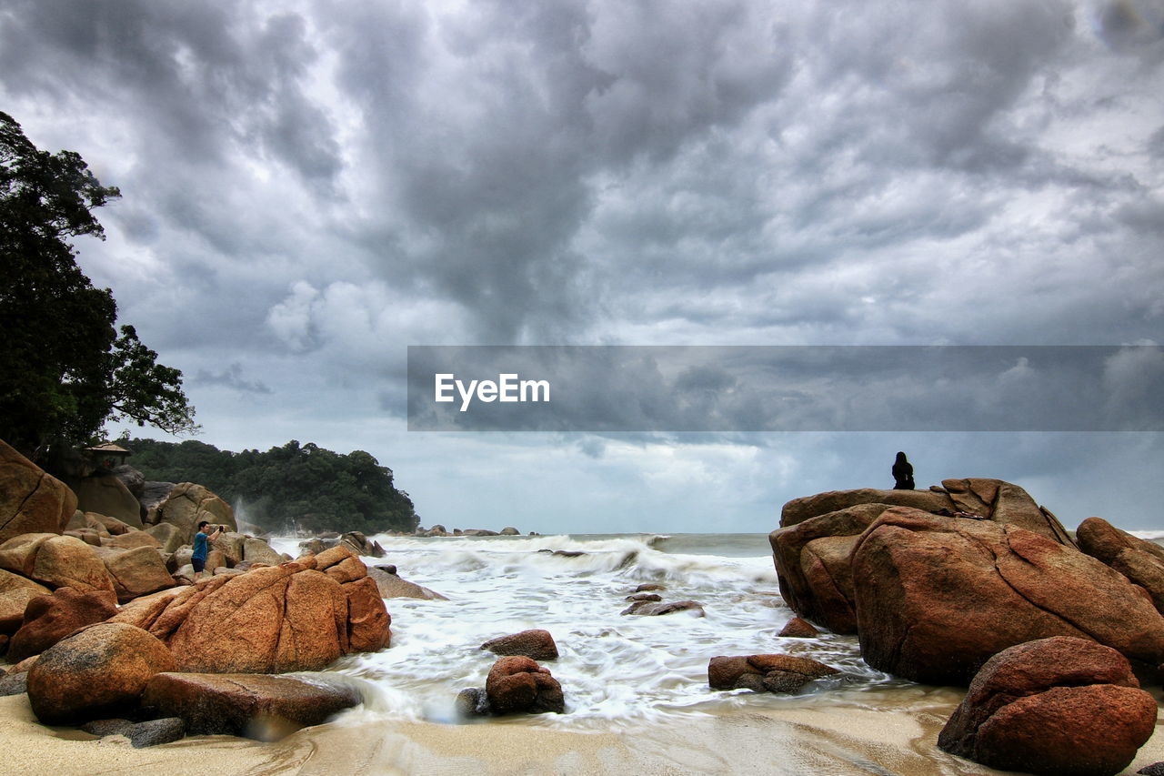 PEOPLE SITTING BY ROCKS ON SEA SHORE AGAINST SKY