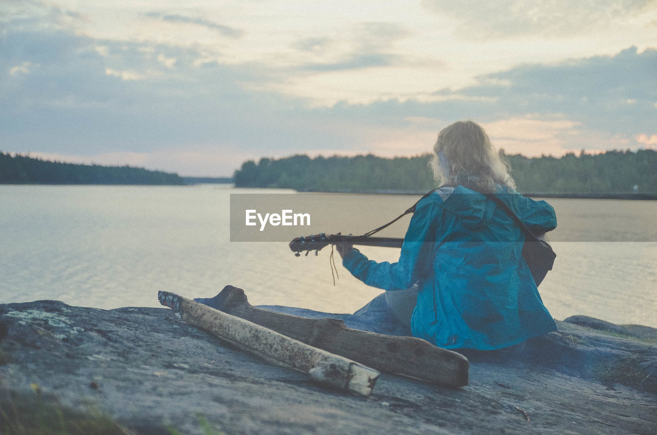 REAR VIEW OF WOMAN SITTING ON SHORE AGAINST SKY