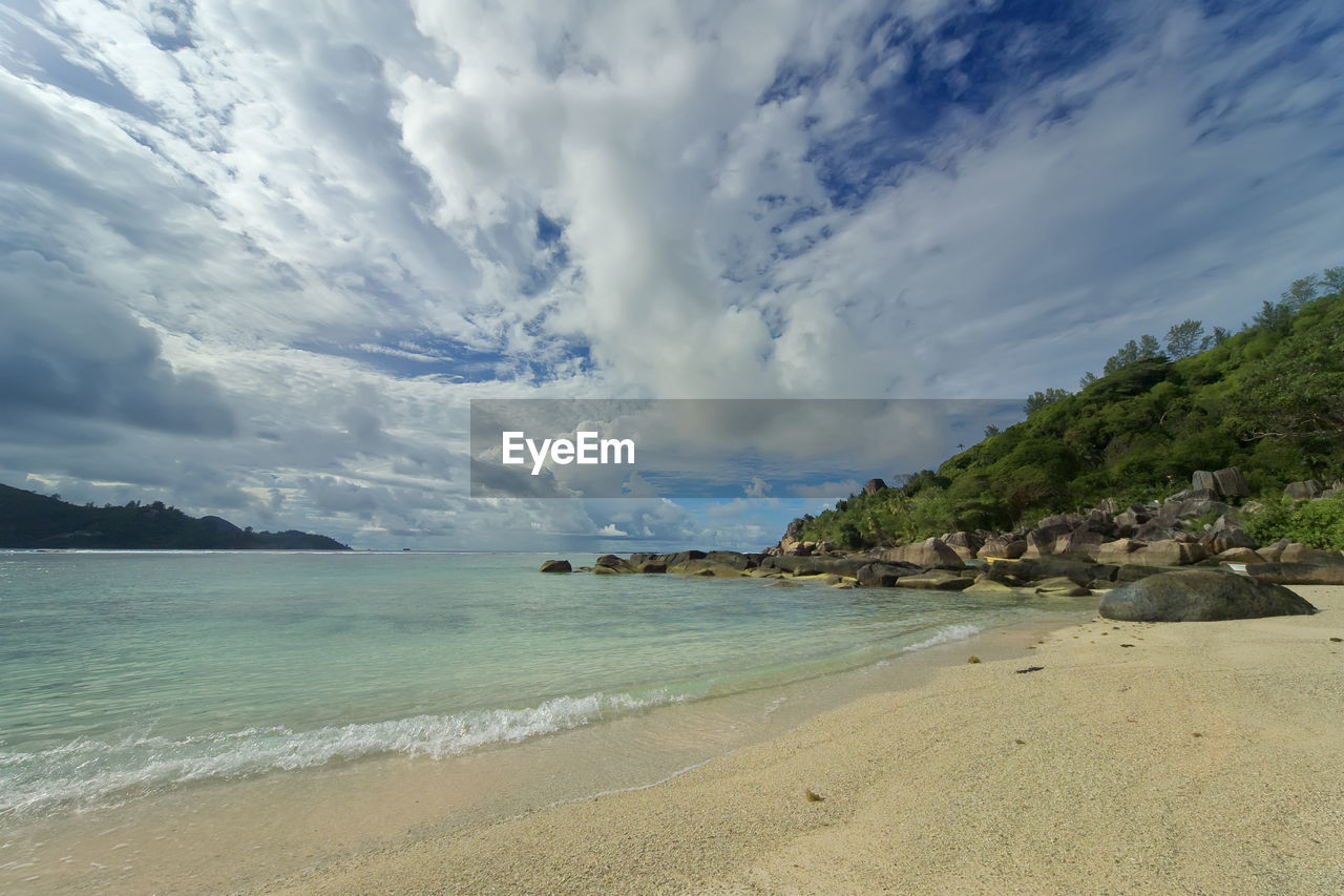 A beach with white and, clear turquoise water, a green coast with boulders going into the water.