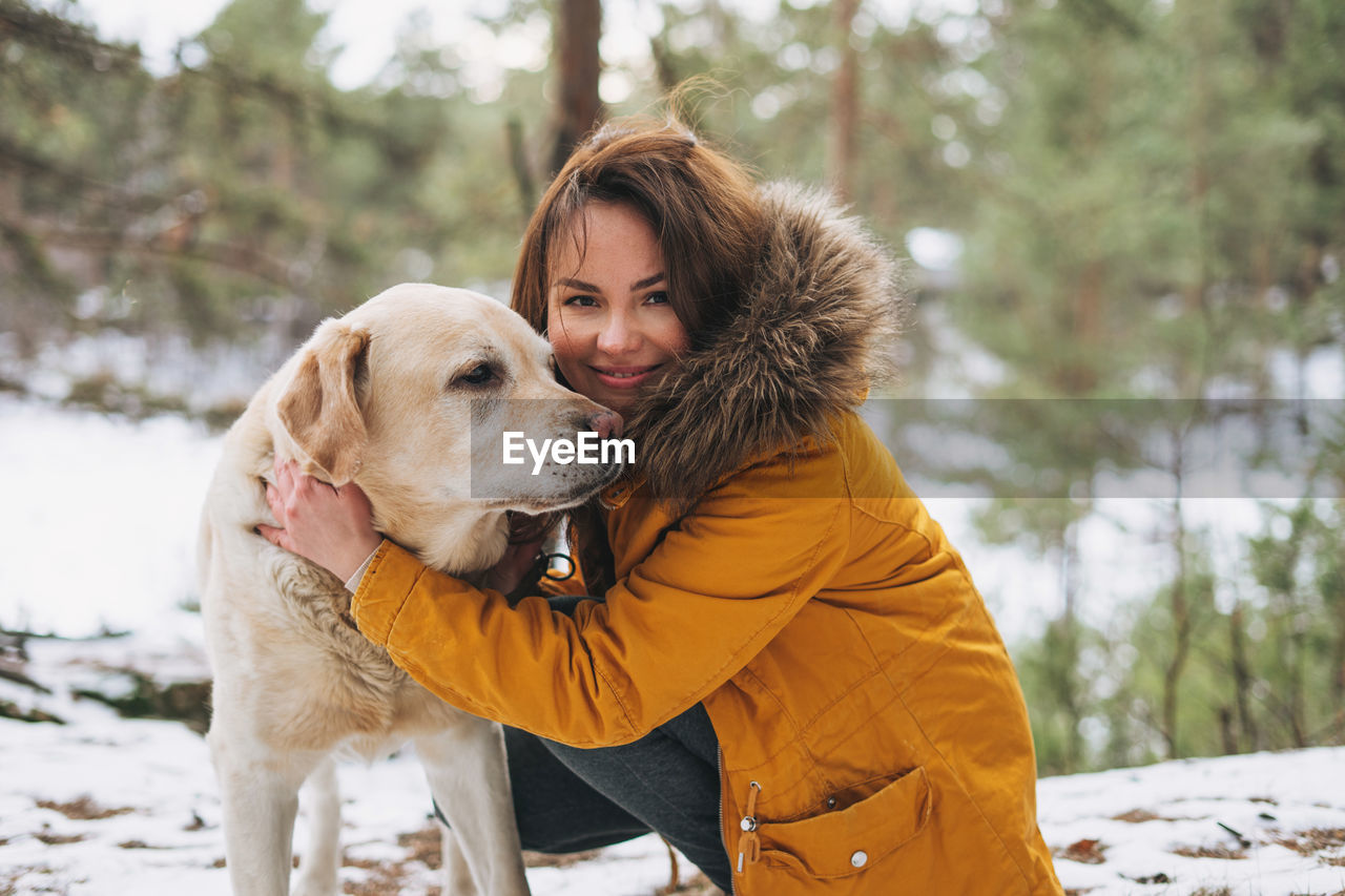 Young smiling woman in yellow jacket with big kind white dog labrador walking in winter forest