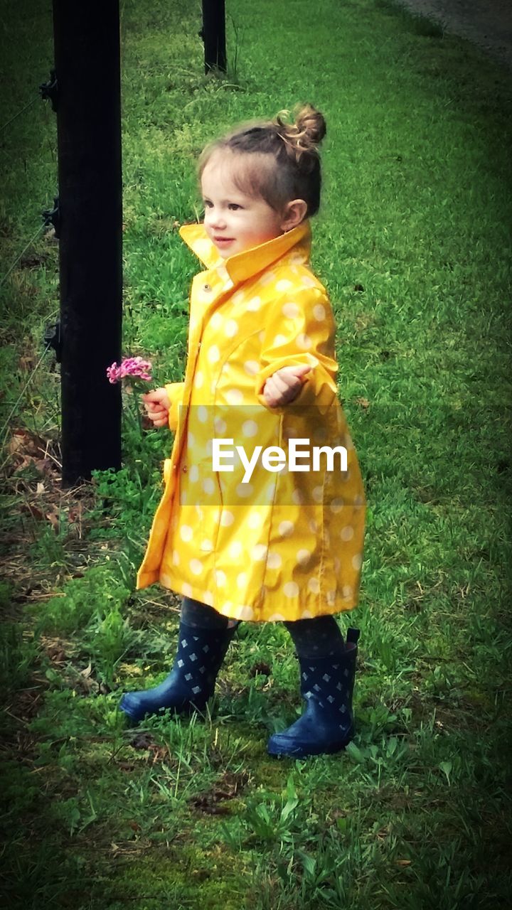 Full length of girl in yellow raincoat holding flower on grassy field