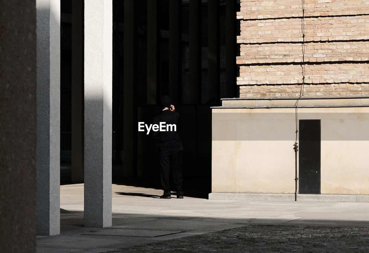 Rear view of man walking by building in city holding a phone