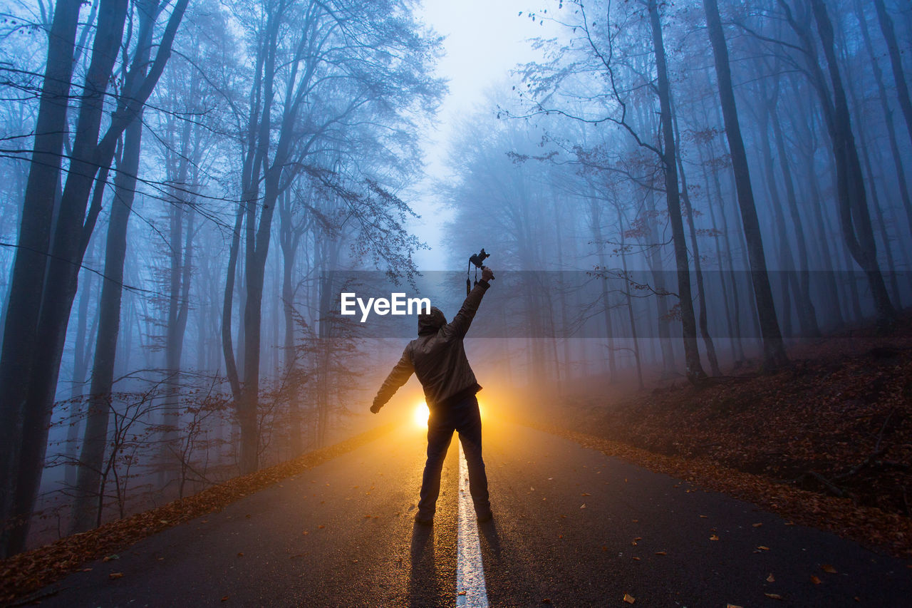 Rear view of man standing on road amidst trees in forest during winter