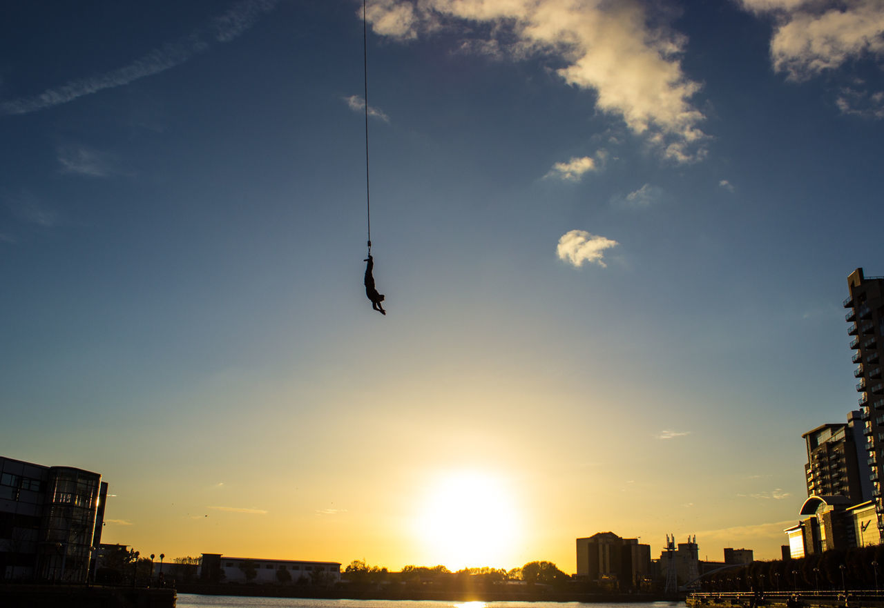 Person bungee jumping over river in city against sky during sunset