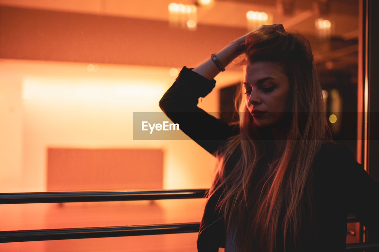 PORTRAIT OF YOUNG WOMAN LOOKING AWAY WHILE STANDING IN ILLUMINATED ROOM