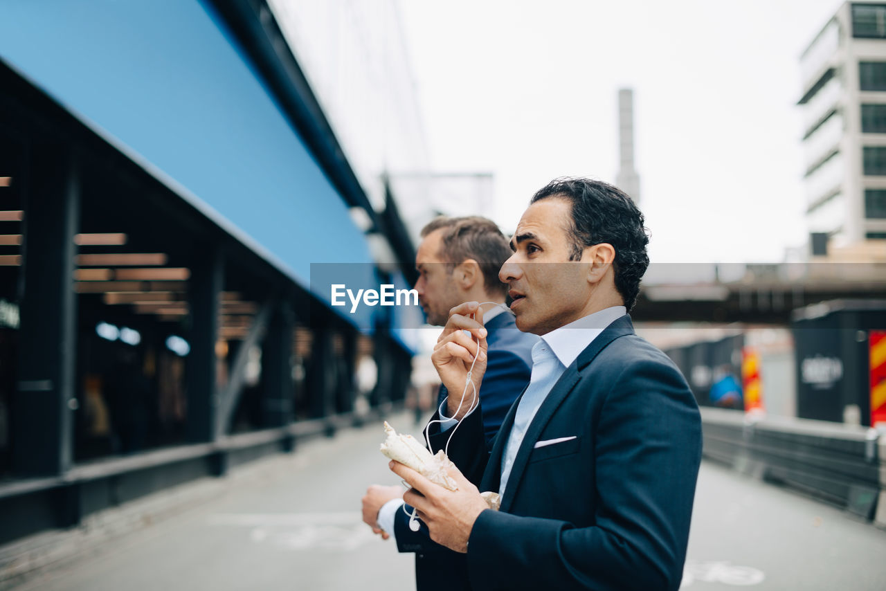 Side view of mature businessman talking through in-ear headphones while standing by male colleague in city