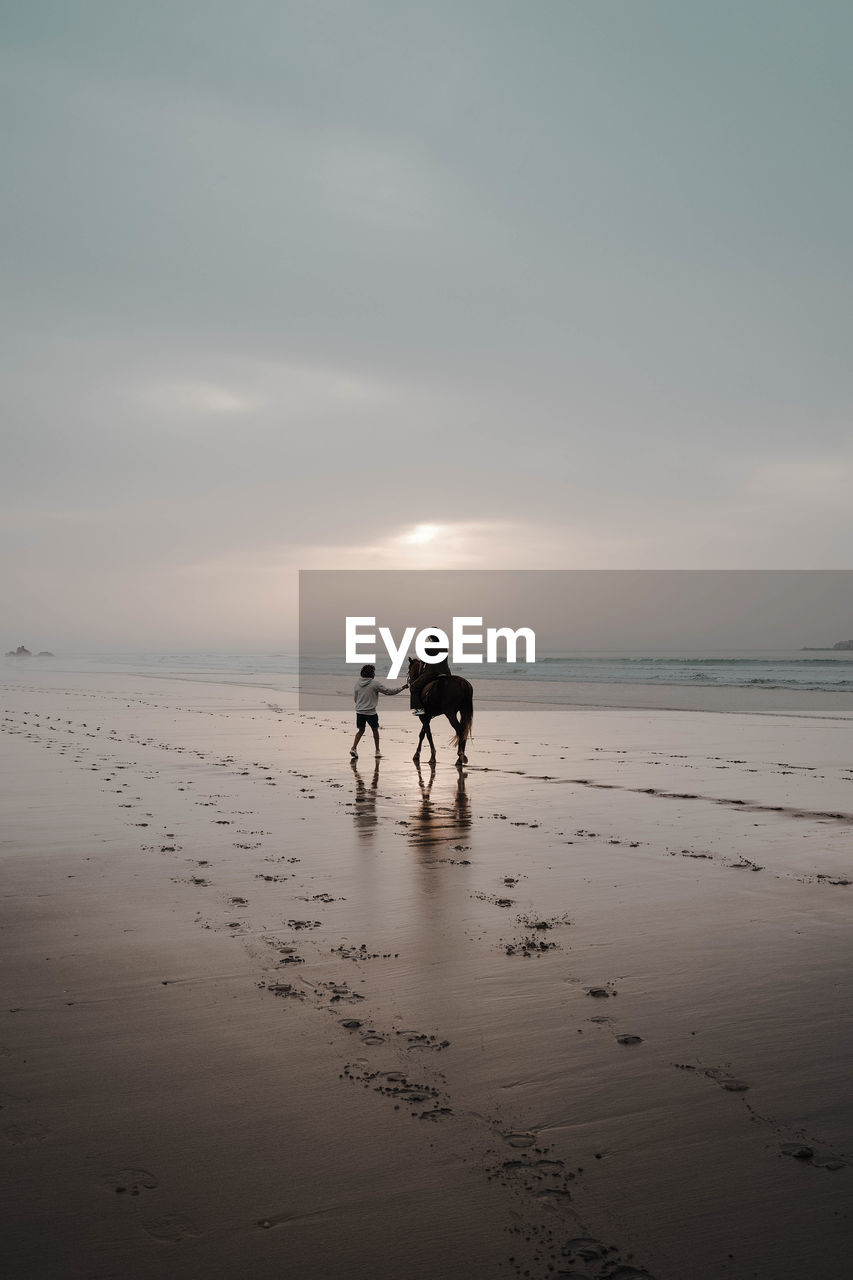 Silhouette woman walking at beach against sky during sunset and a person riding a horse