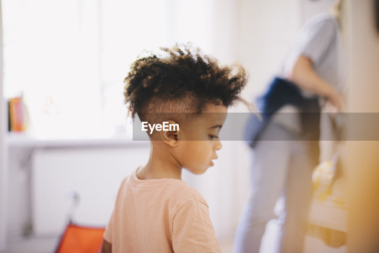 Boy with curly hair looking down while mother working in background