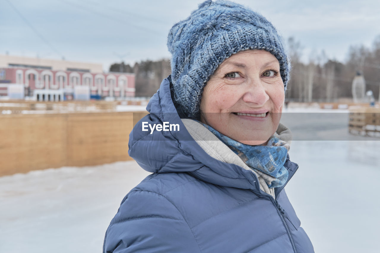 Senior white smiling woman  in blue jacket and knitted hat, on skating rink in winter. 