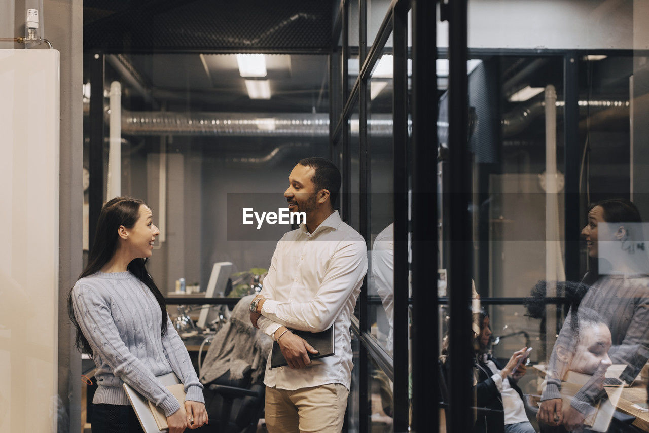 Smiling male and female professional colleagues talking while standing at illuminated office