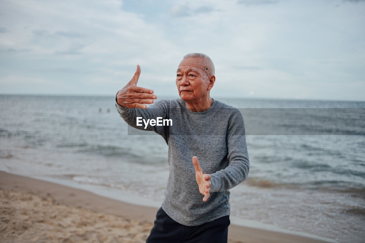 Senior man exercising while standing at beach against cloudy sky during sunset