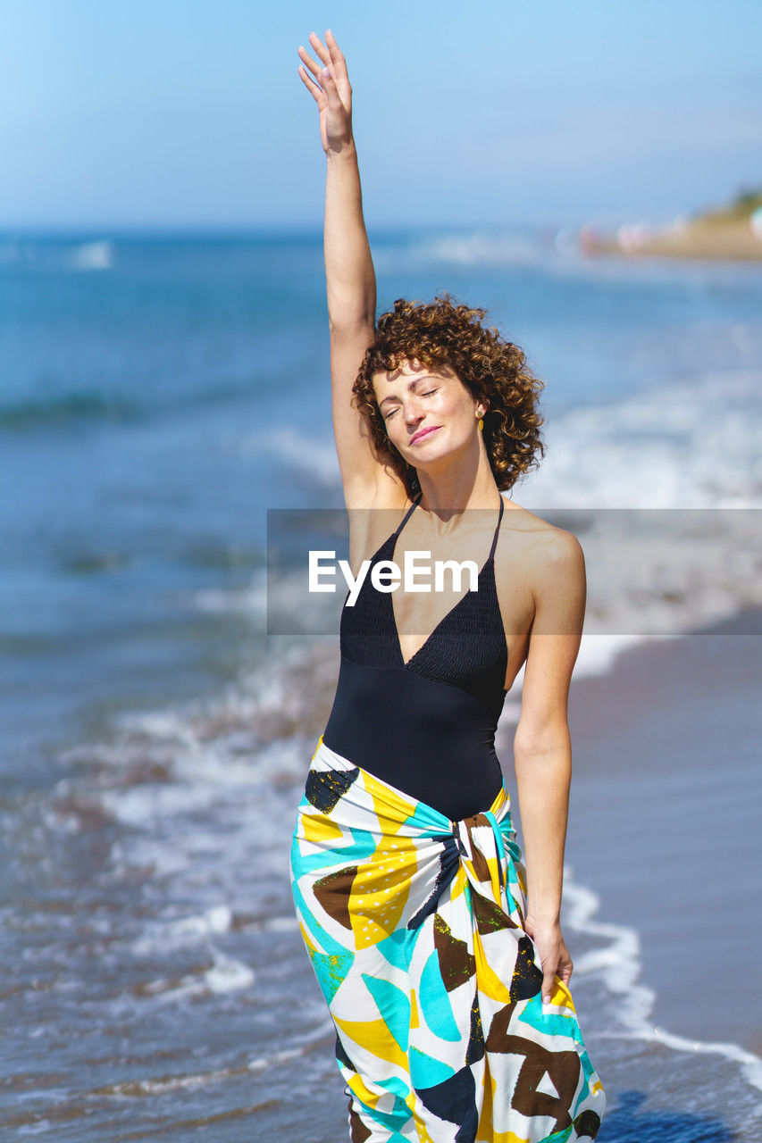 portrait of young woman with arms raised standing at beach