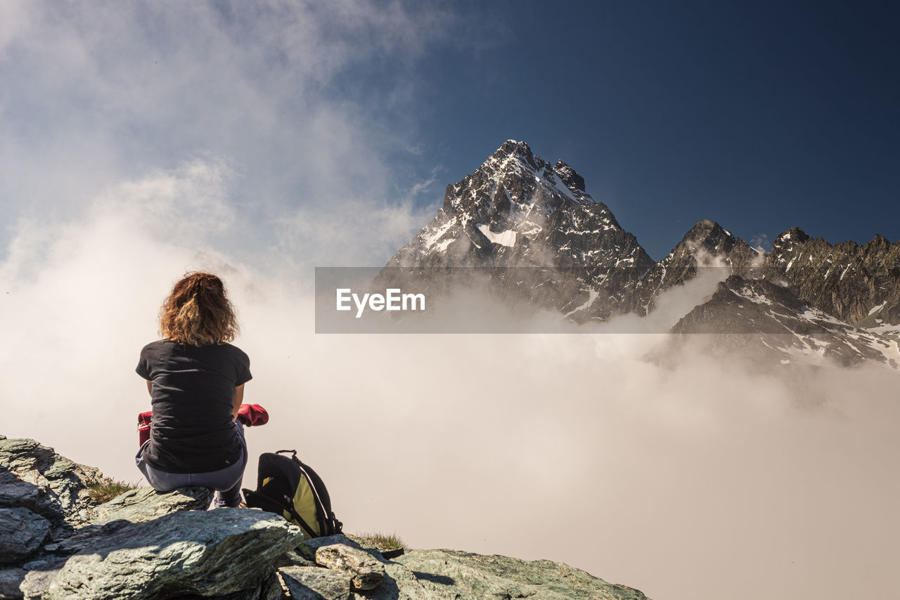 REAR VIEW OF WOMAN SITTING ON ROCK AGAINST MOUNTAIN