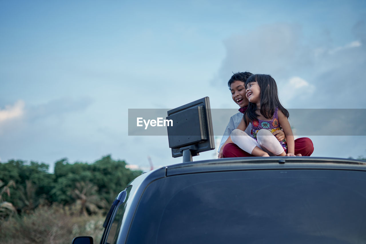 Cheerful siblings looking at desktop pc while sitting on car