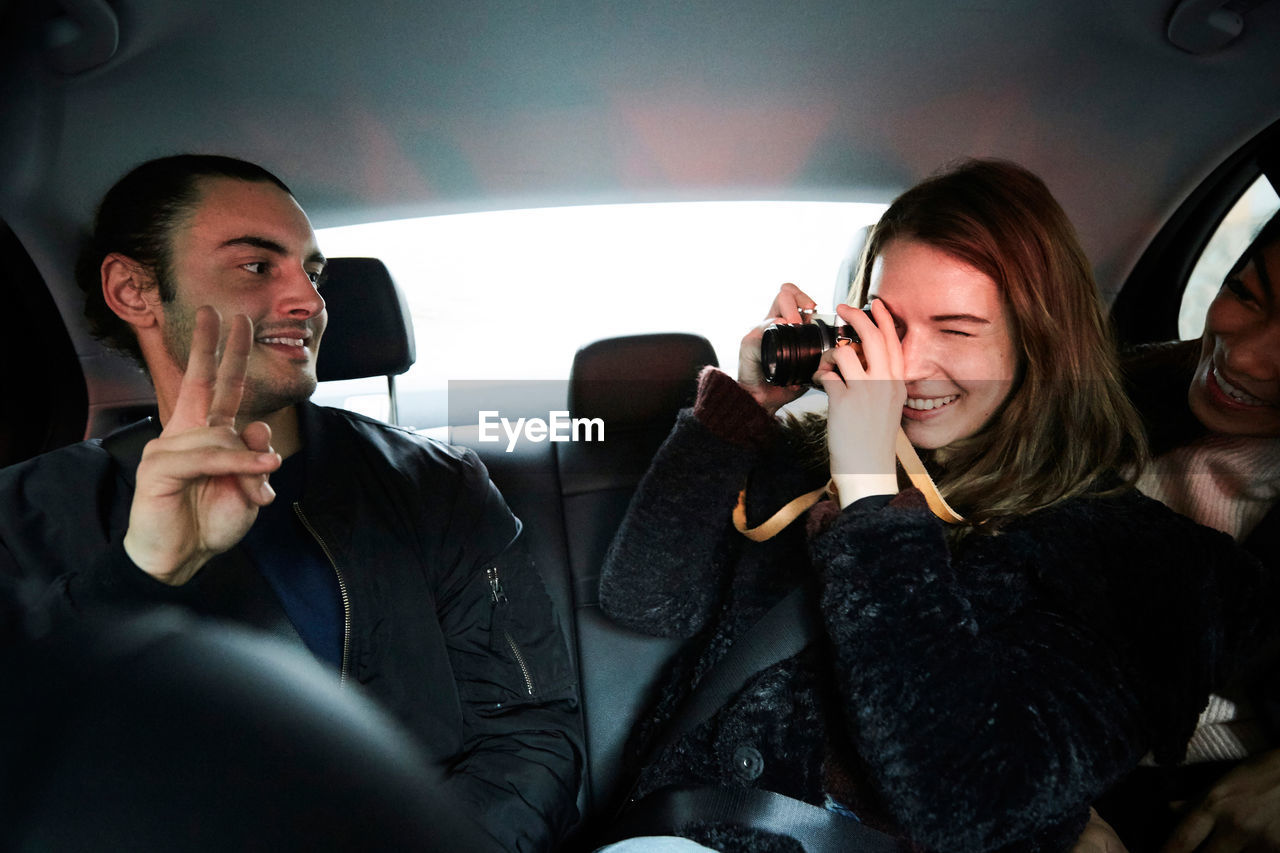 Smiling woman photographing male friend gesturing peace sign while riding in taxi at city