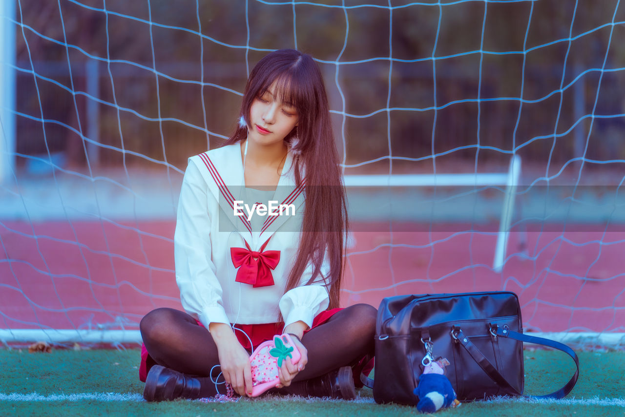 Young woman listening to music while sitting against net on soccer field