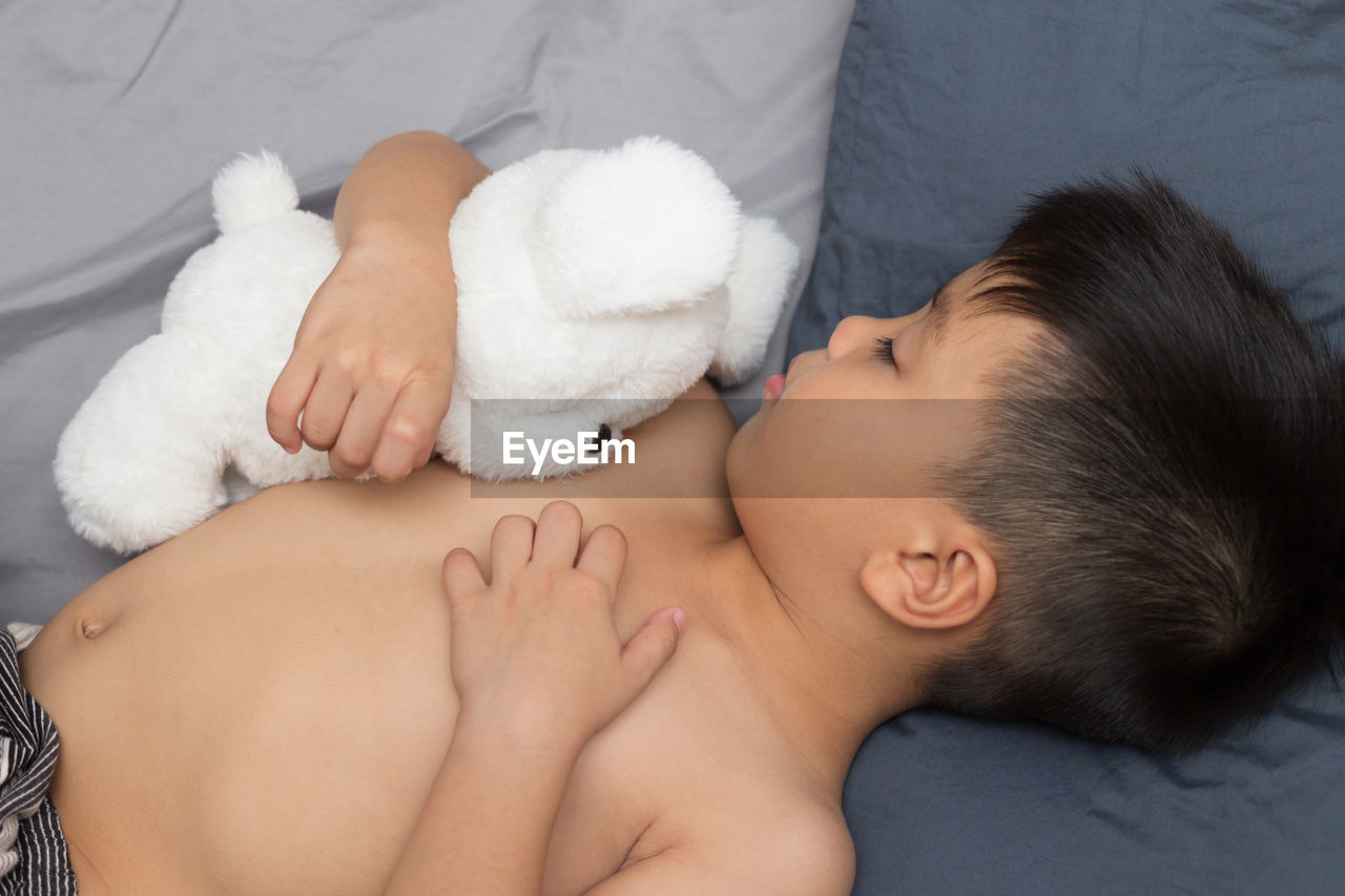 High angle view of shirtless boy with stuffed toy sleeping on bed