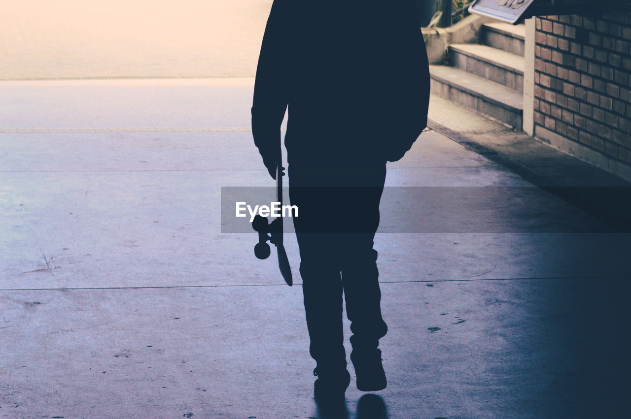 Low section of silhouette man carrying skateboard on footpath