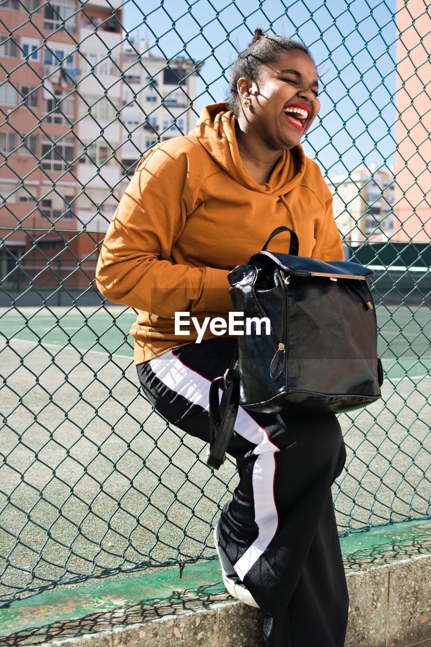 Smiling young woman with bag leaning on chainlink fence