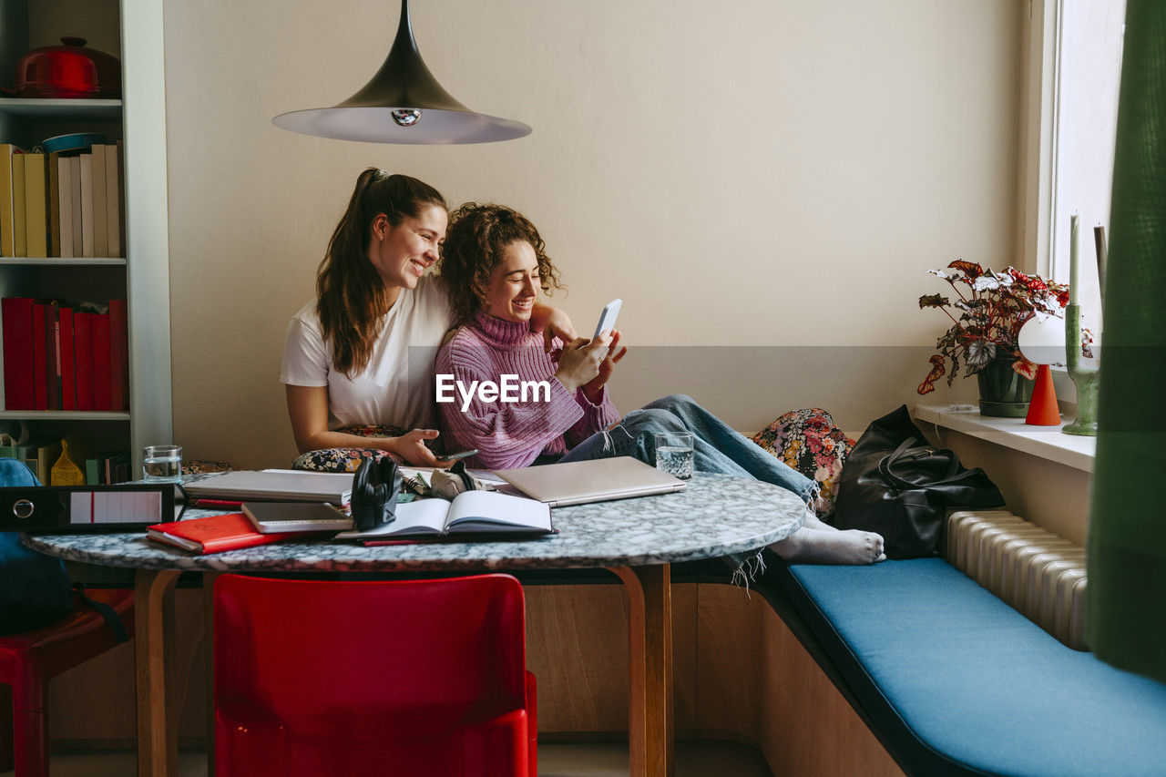 Happy young female friends sharing smart phone while sitting at home