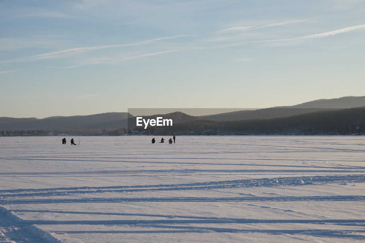 Fishermen sit and fish on the snow-covered lake in the evening light.