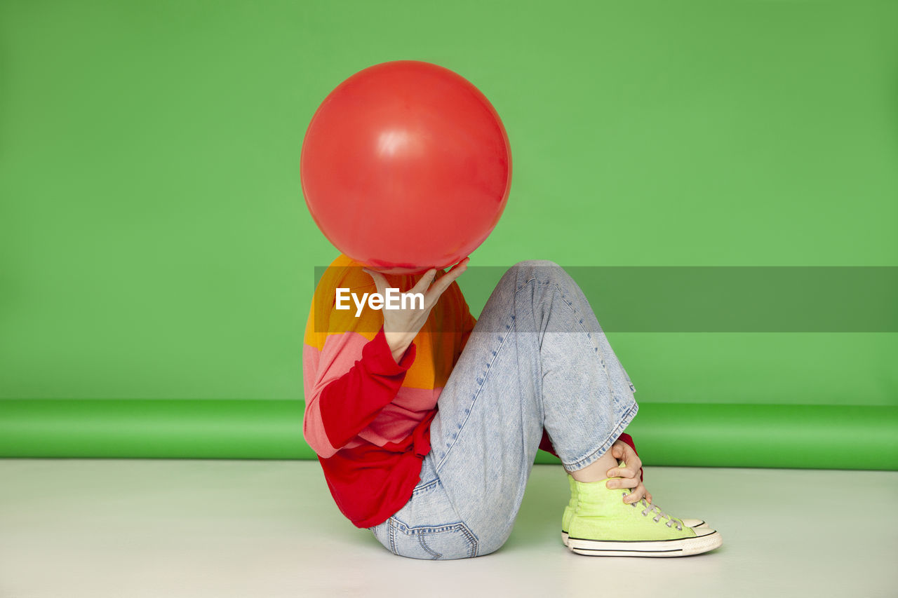 Woman covering face with red balloon sitting against green background