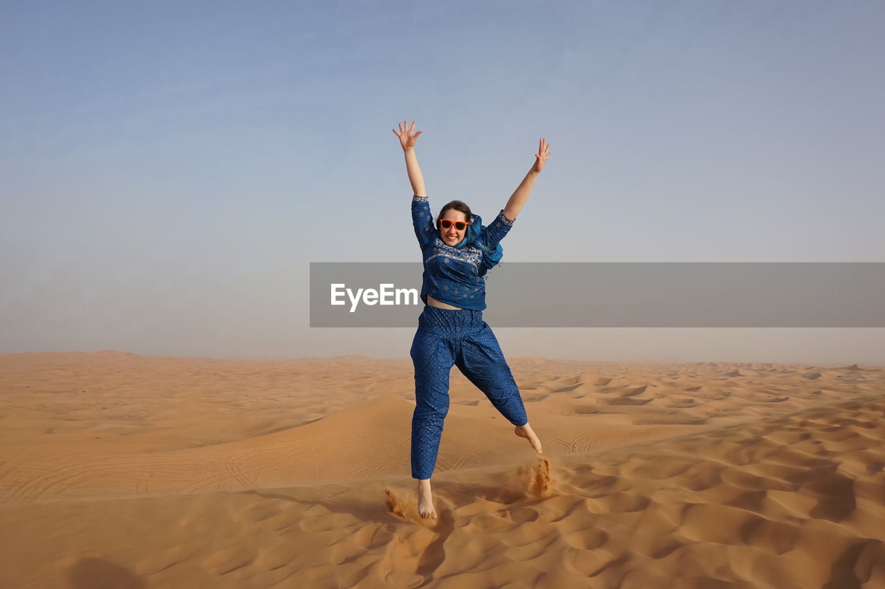 Full length of excited woman jumping at desert against clear sky