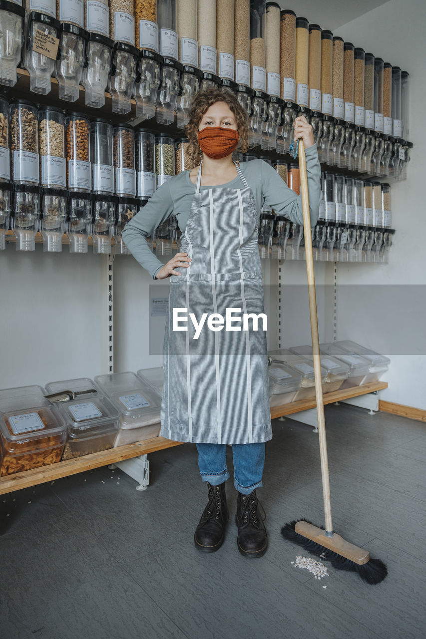 Female store clerk with hand on hip holding broom while standing in zero waste shop during covid-19