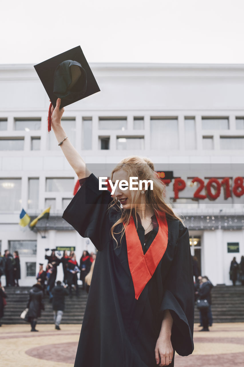 Woman in graduation gown holding mortarboard while standing against building