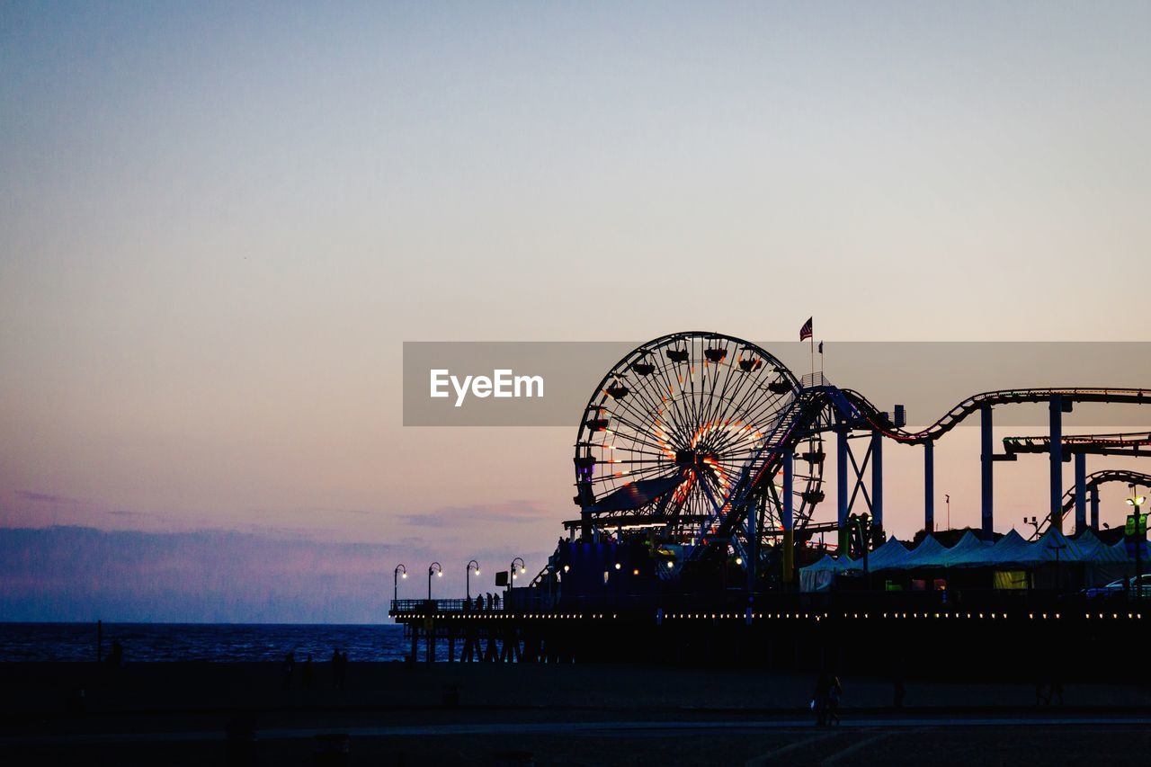 Illuminated amusement park by sea against sky