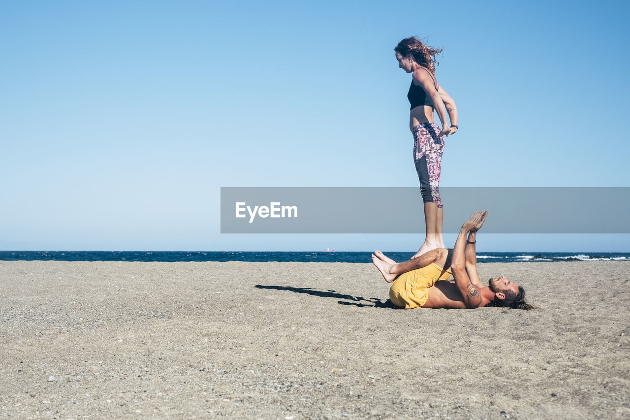 Couple practicing yoga at beach against clear blue sky