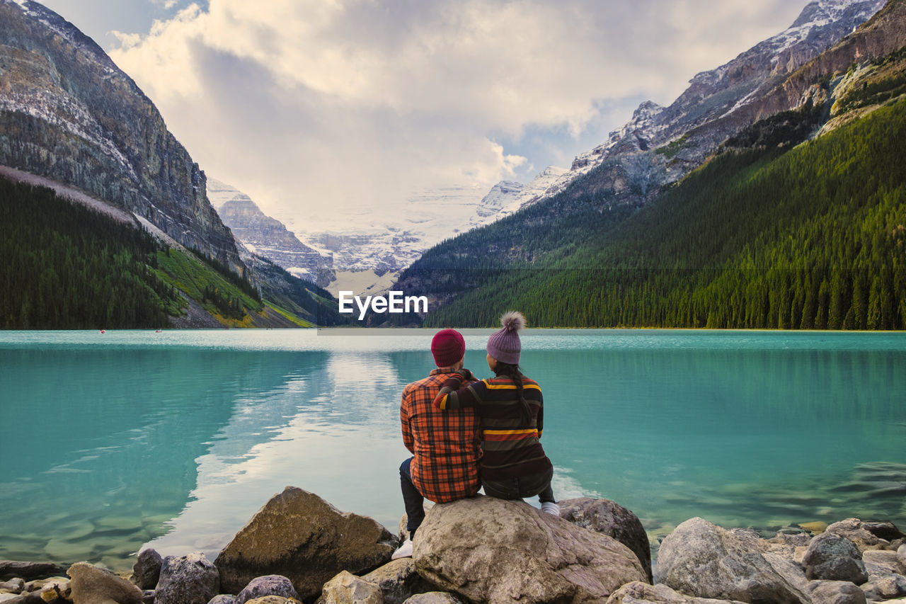 rear view of man sitting on rock by lake by snowcapped mountains against sky