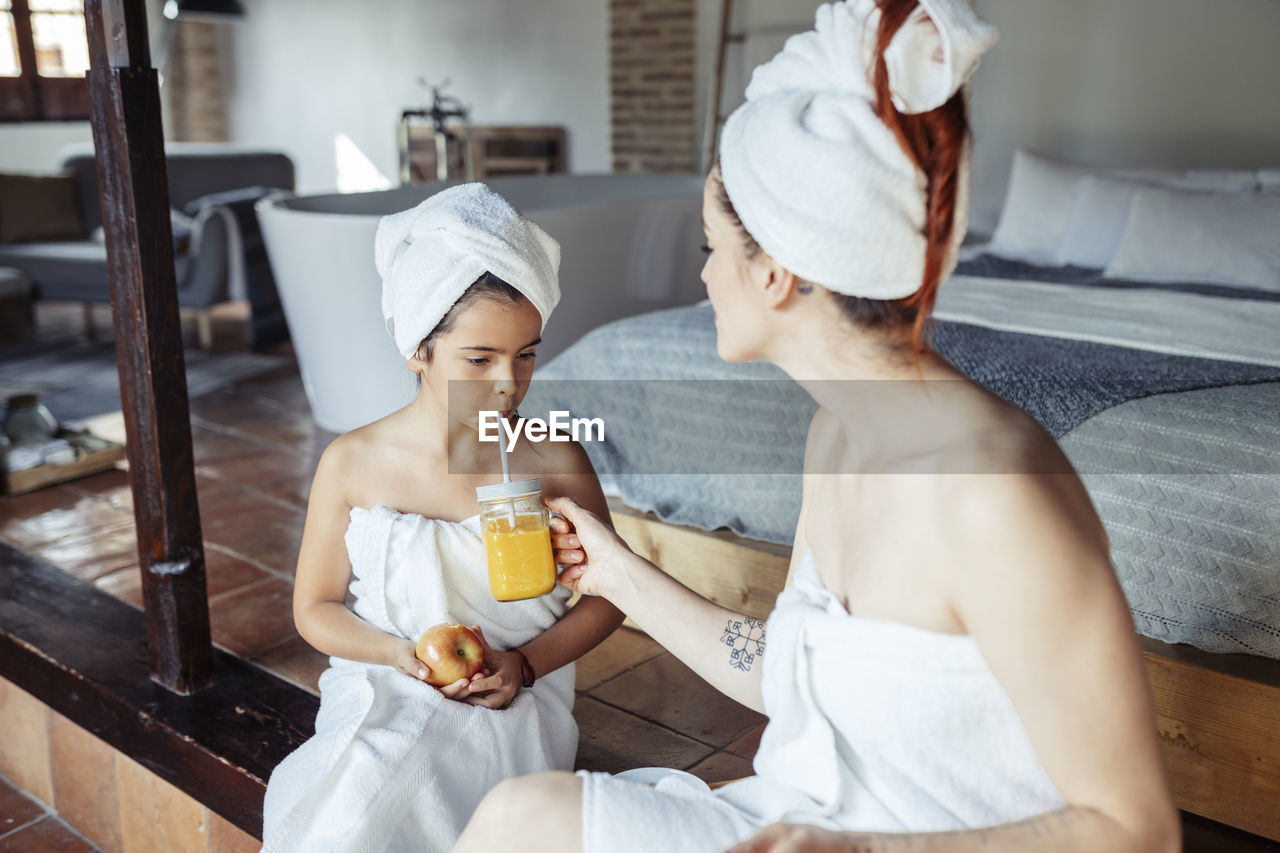 Mother in towel feeding juice to daughter while sitting at home