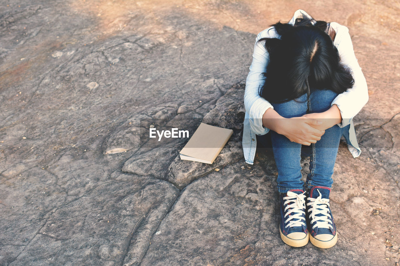 High angle view of woman hugging knees while sitting by book on rock