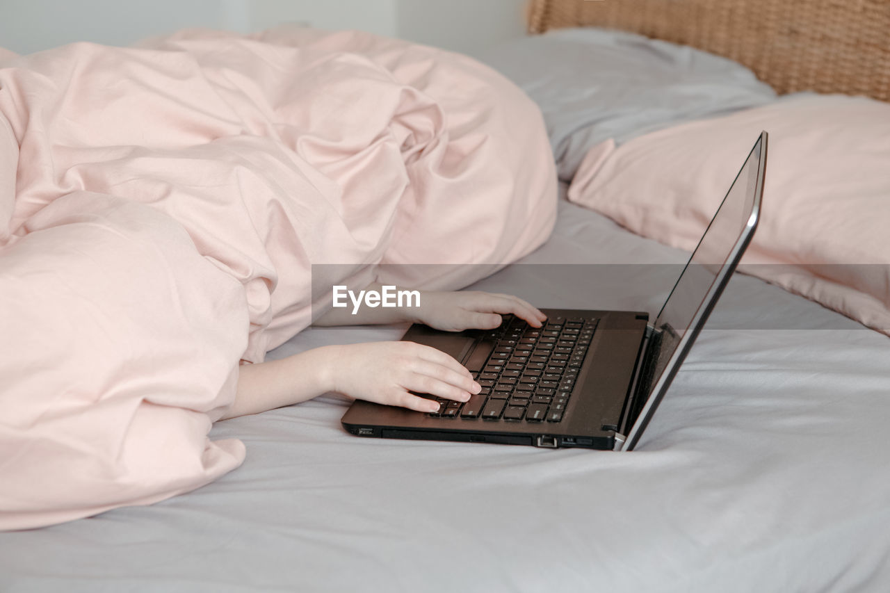 Bored funny child lying in bed under blanket and learning in virtual online school class. hands 
