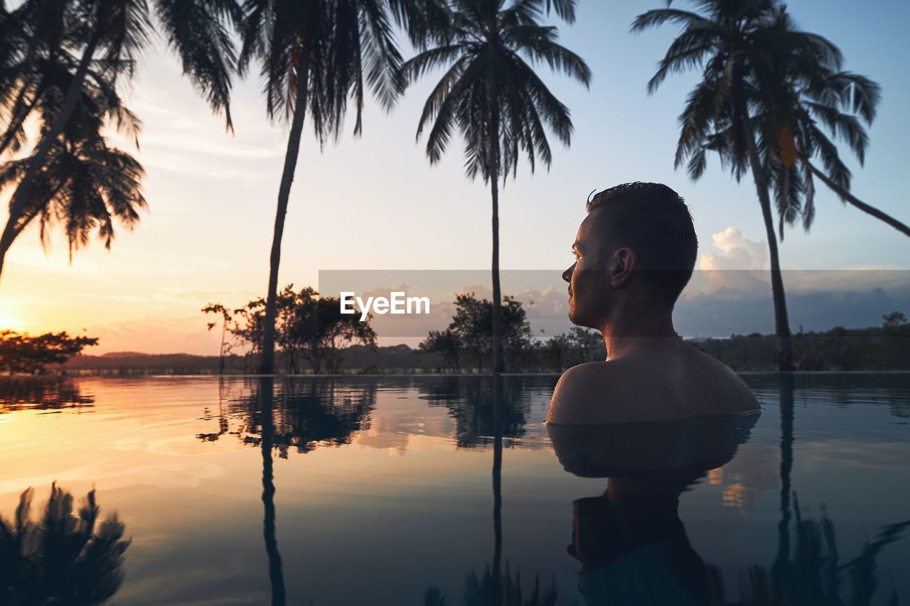 Young man watching sunset from swimming pool in the middle of coconut palm trees.