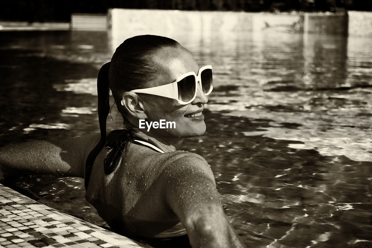 A vintage photo of a woman in a black swimsuit and white sunglasses swimming in the pool