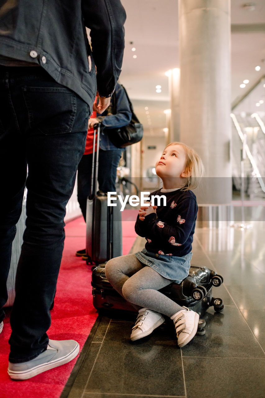 Full length of girl with hands clasped looking at father while sitting on suitcase in hotel
