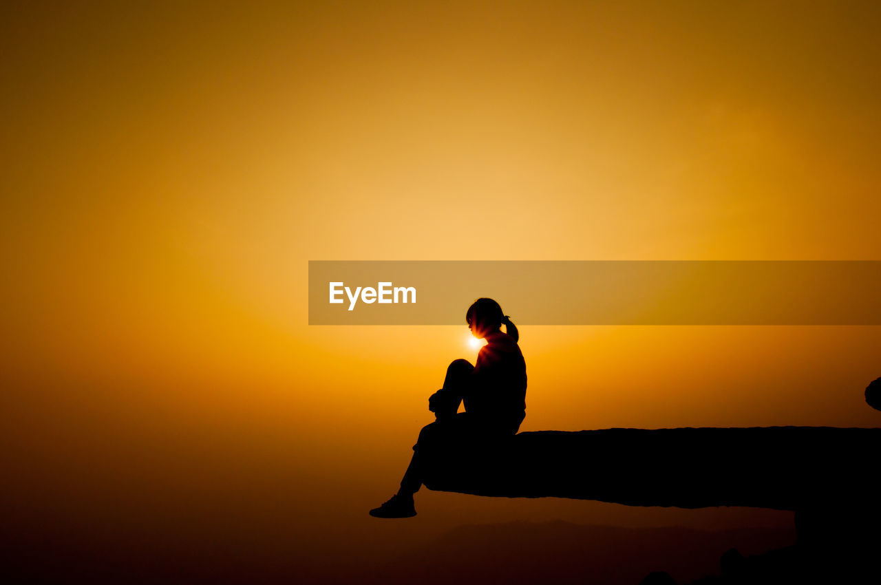 Silhouette of woman sitting on cliff against sky during sunset