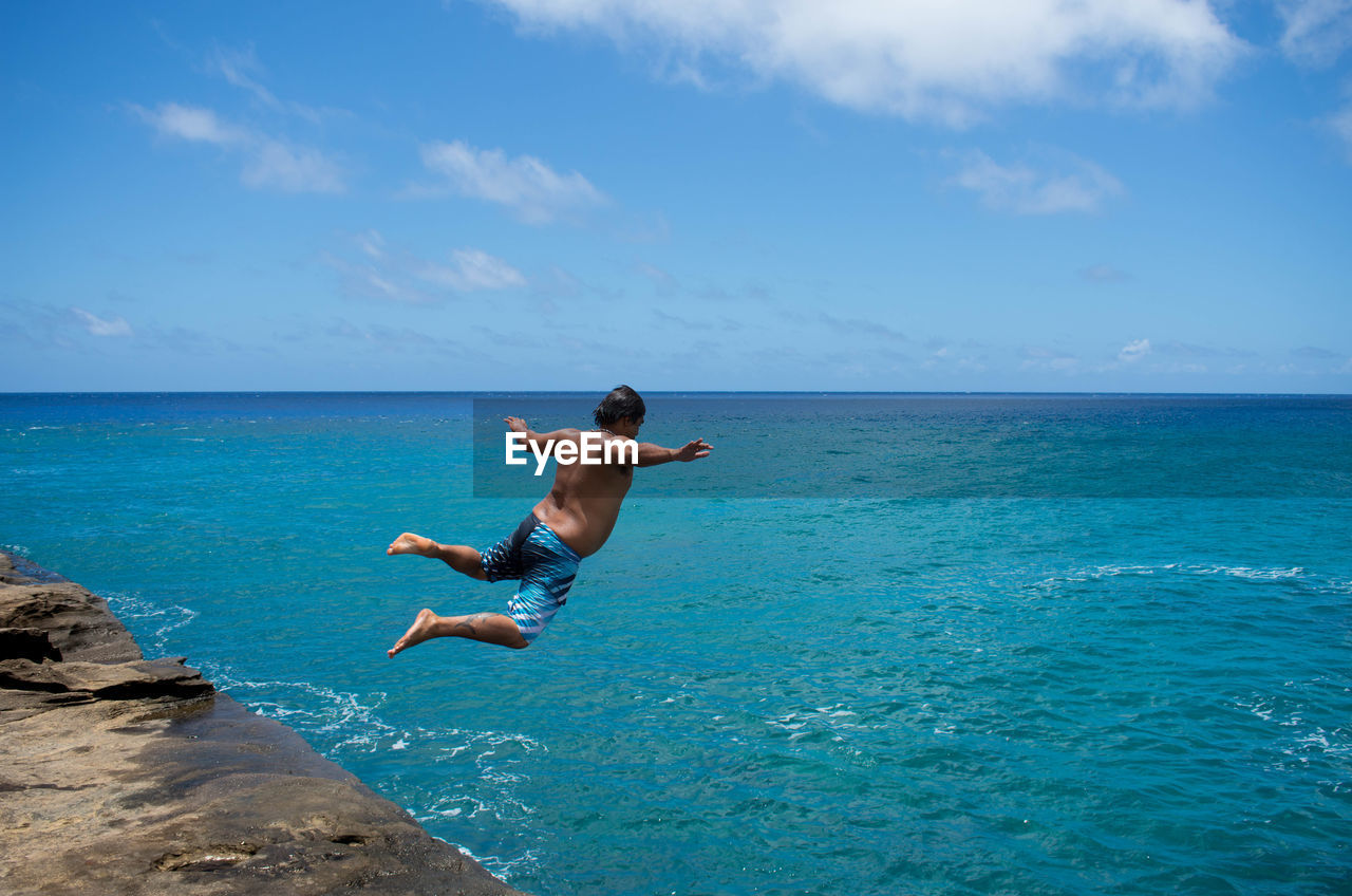REAR VIEW OF MAN JUMPING IN SEA AGAINST SKY