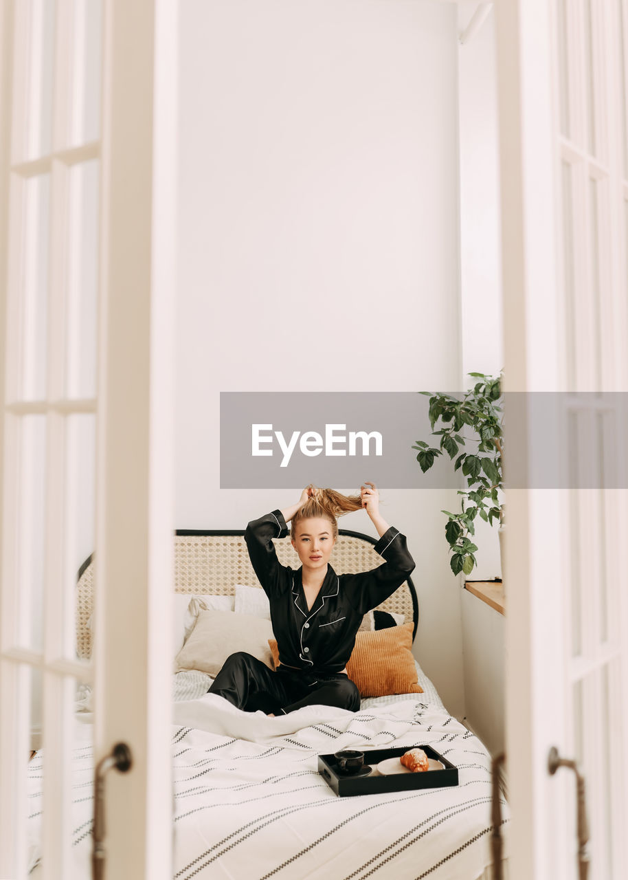 A happy pretty young woman in pajamas is resting and having breakfast in bed in a cozy apartment