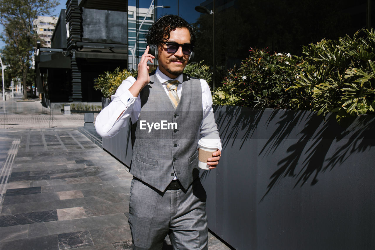 Positive bearded male entrepreneur in elegant suit having phone conversation on smartphone and looking away while standing with takeaway coffee on the street with sunglasses