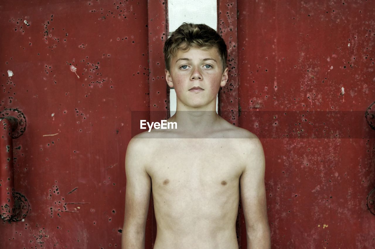 Portrait of shirtless teenage boy standing against red gate