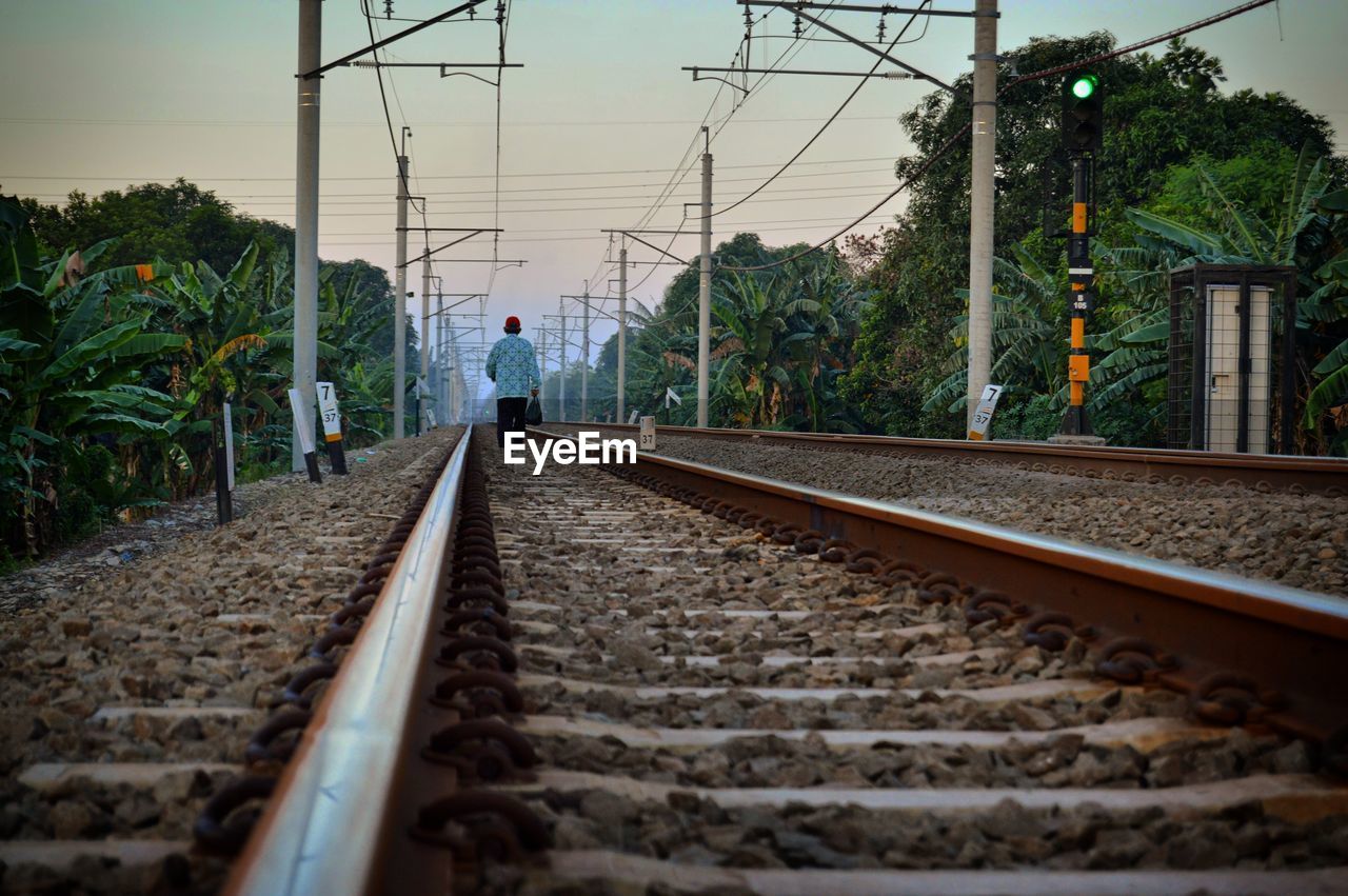 Rear view of man walking on railroad tracks against sky during sunset