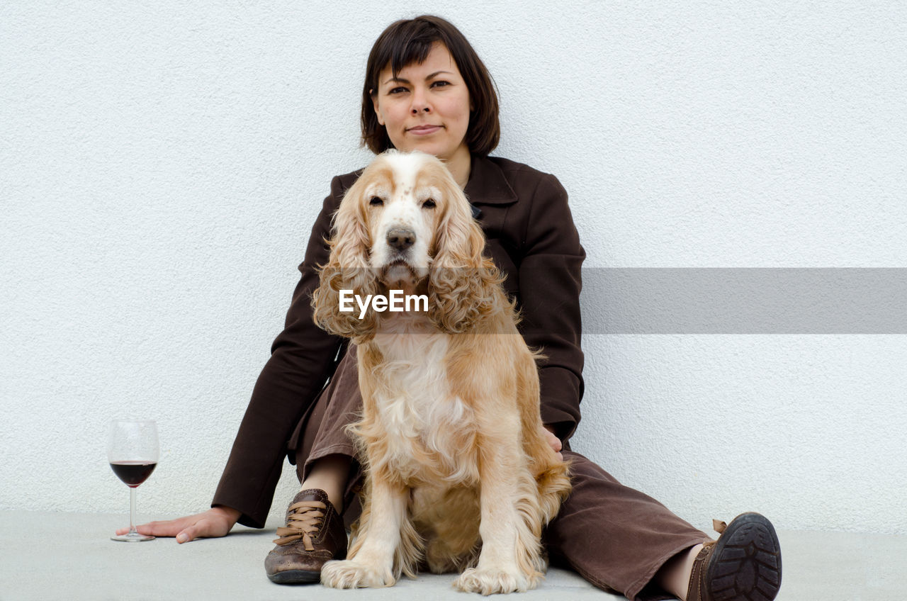 Portrait of woman with cocker spaniel by wineglass sitting against wall