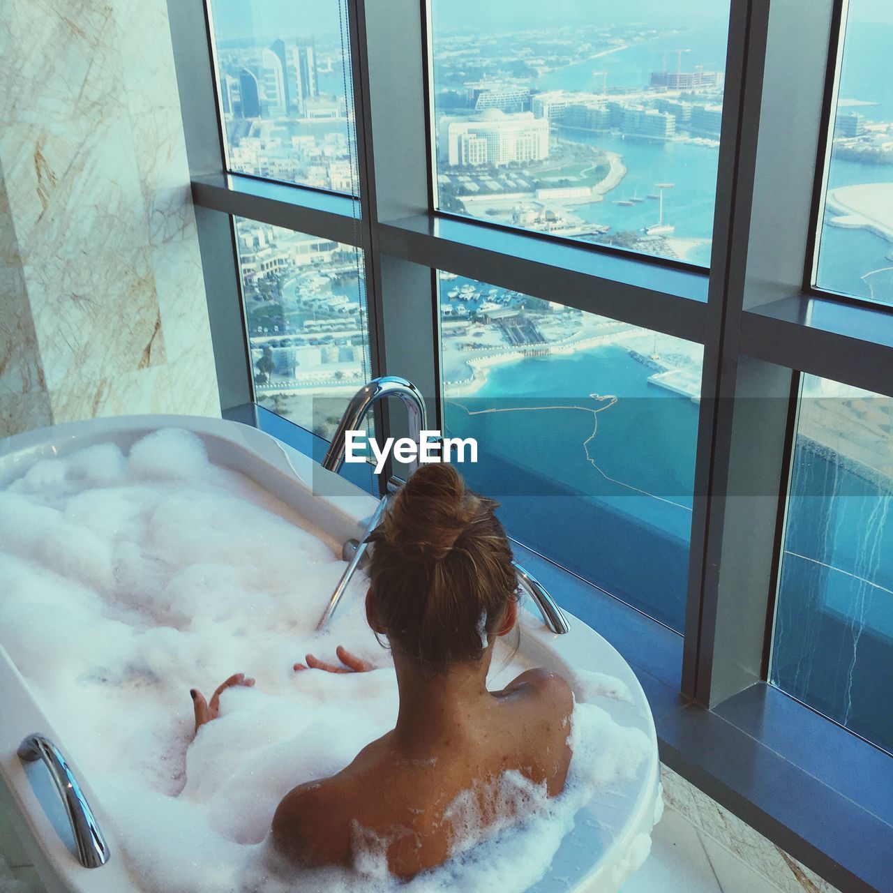 High angle view of young woman sitting in bathtub by window at bathroom