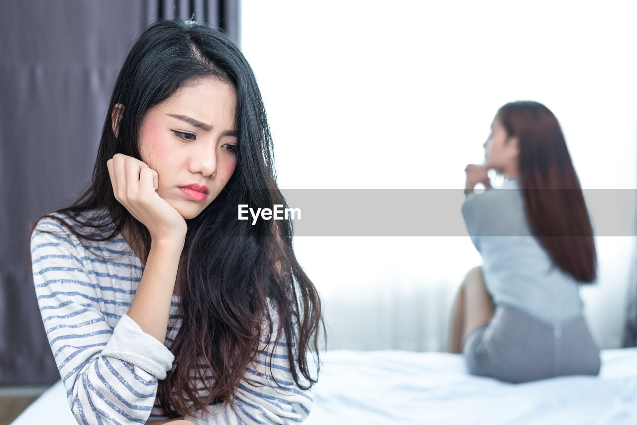 Sad young woman with friend sitting on bed in background