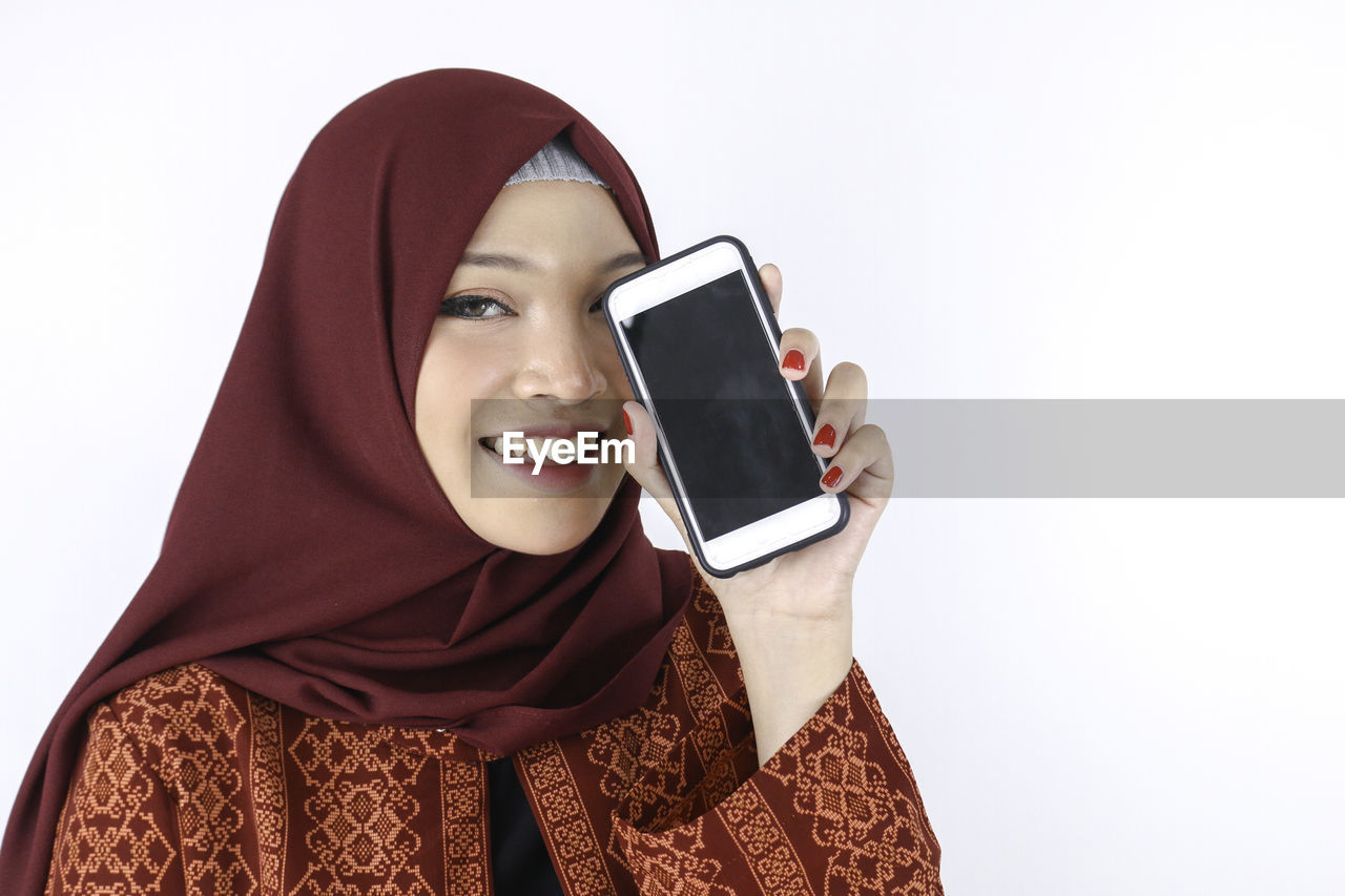 PORTRAIT OF SMILING YOUNG WOMAN USING SMART PHONE AGAINST WHITE BACKGROUND