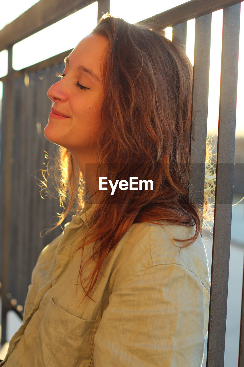 one person, women, adult, hairstyle, young adult, long hair, looking, human hair, lifestyles, smiling, portrait, brown hair, blond hair, headshot, happiness, leisure activity, architecture, casual clothing, window, looking away, female, sunlight, day, nature, outdoors, person, standing, emotion, contemplation, side view, waist up, human face, enjoyment, clothing, relaxation, cheerful, carefree, redhead