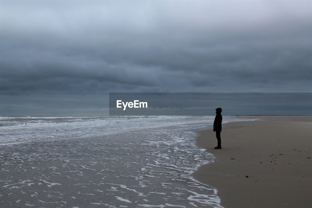 Side view of man standing on shore at beach against cloudy sky